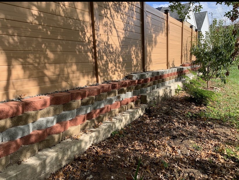 Multi-tone stacked block retaining wall running along a wood-tone fence line